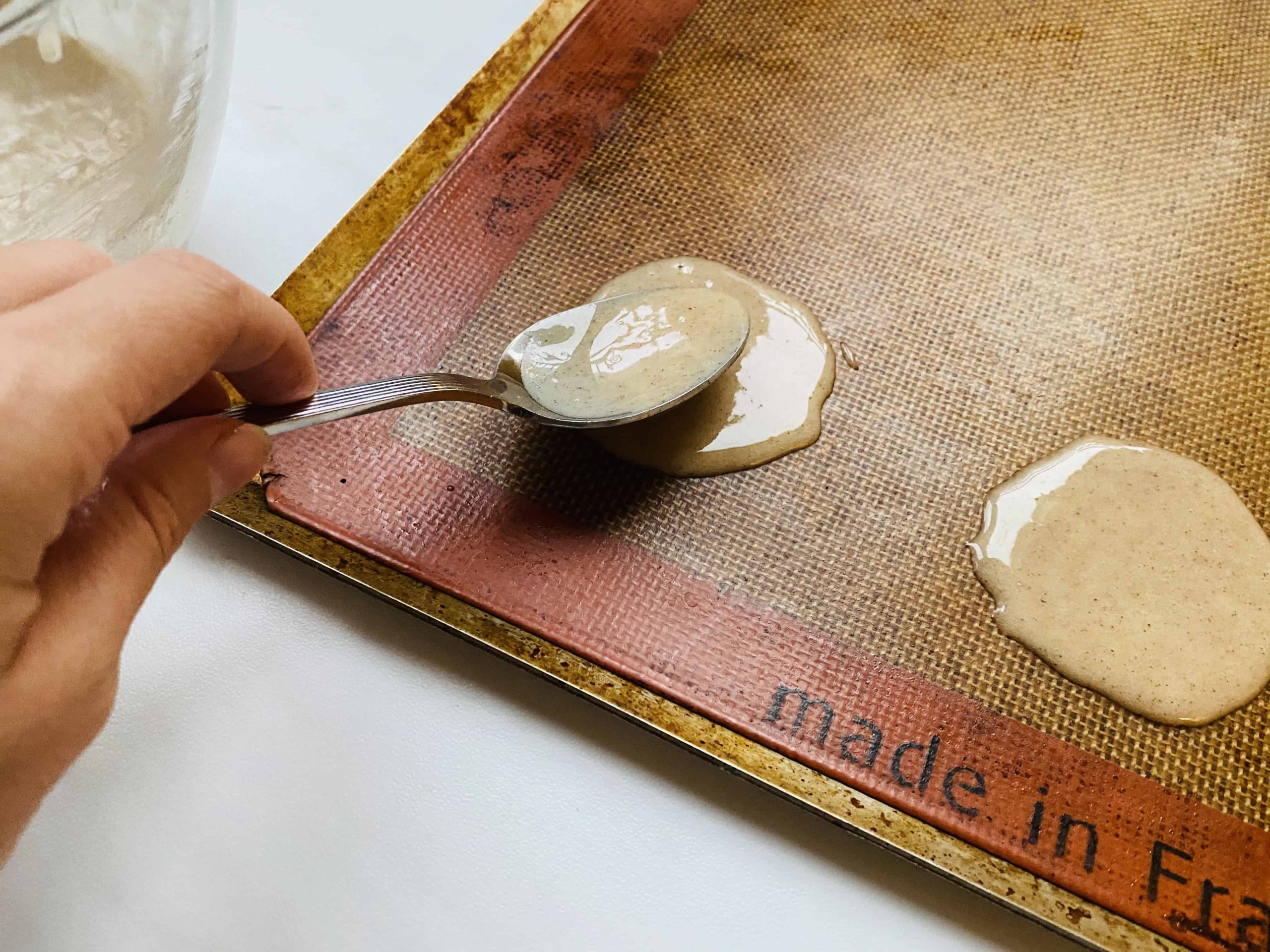 A hand spreading out cookie batter with a spoon on a baking sheet.