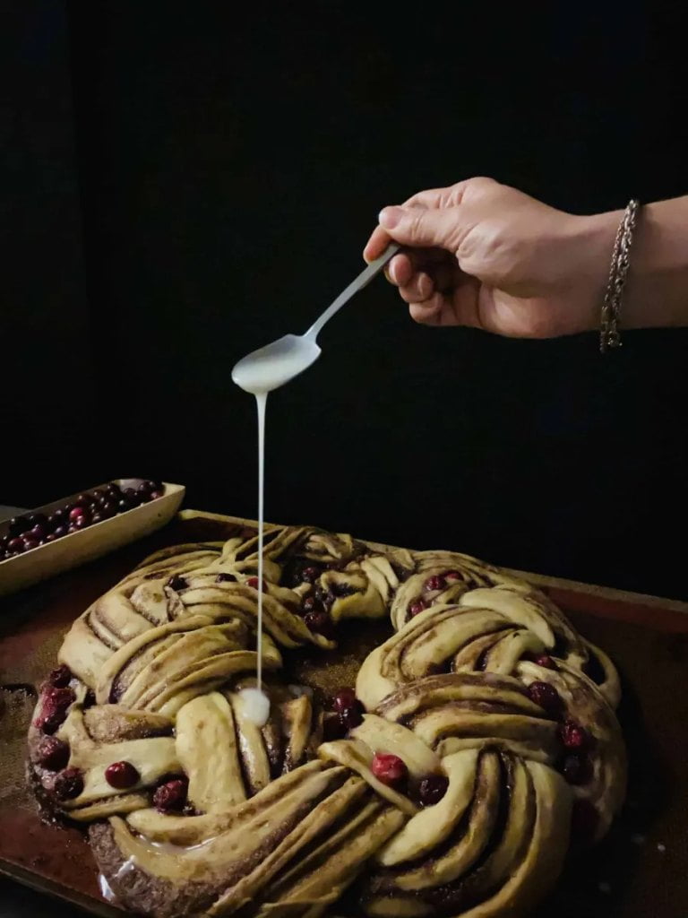 A hand drizzling icing from a spoon onto a wreath of baked cinnamon roll dough.