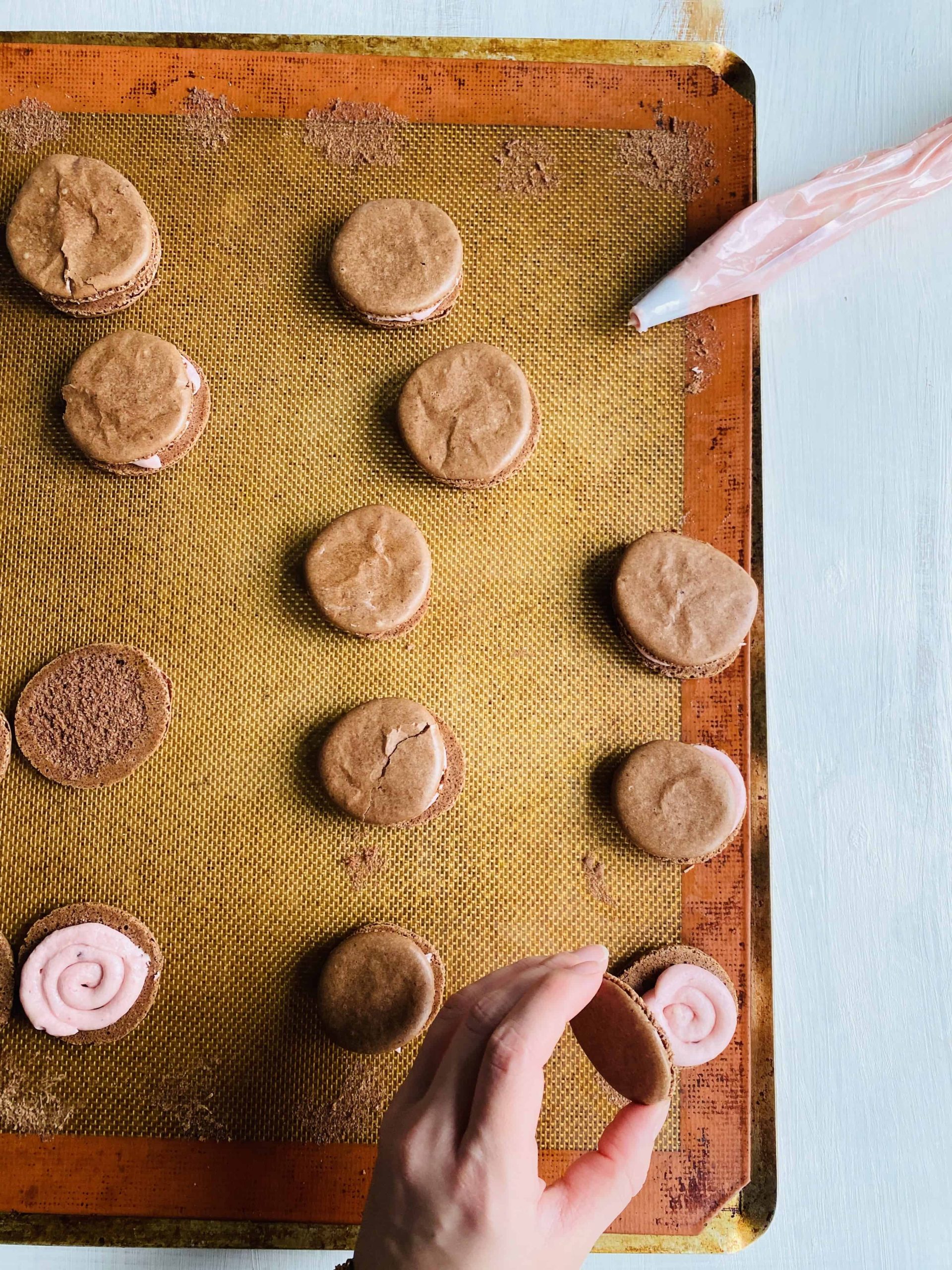 Chocolate macarons being piped with swirls of strawberry buttercream.
