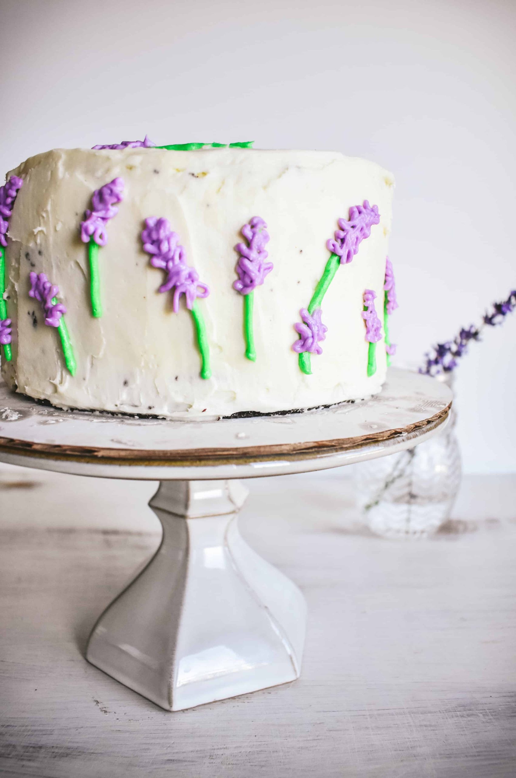 A four-layer cake decorated with lavender flowers piped with buttercream icing on a cake stand.