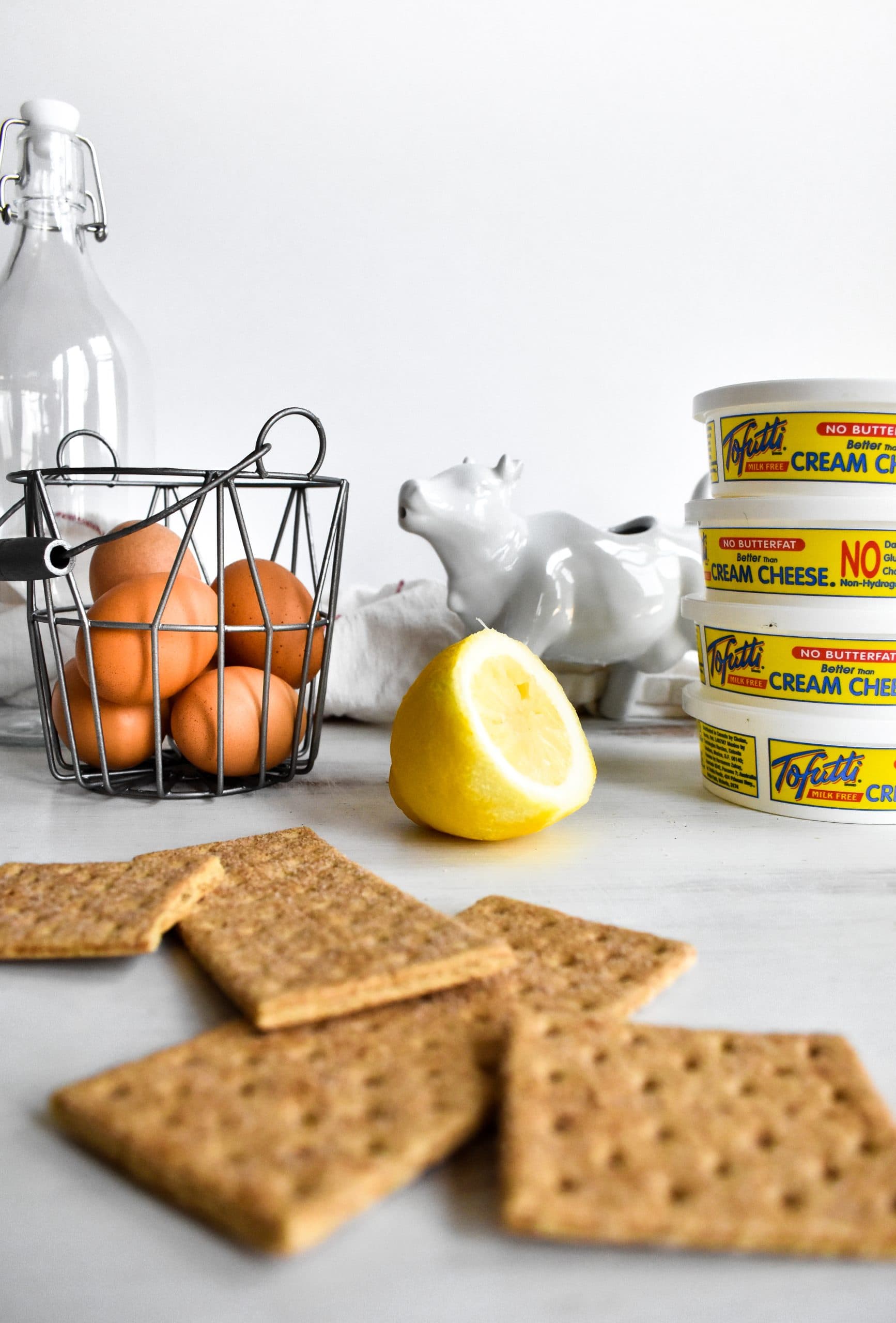 Graham crackers, half a lemon, eggs in a basket, and tubs of dairy-free cream cheese laid out on a light table.