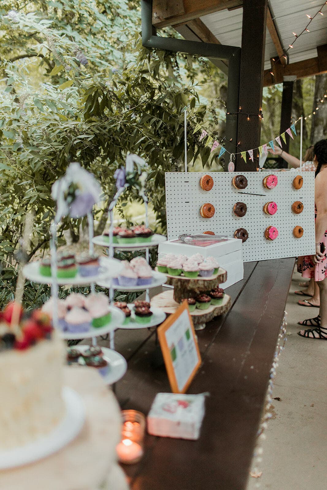 A wedding dessert table with cake, cupcakes and a donut wall in a wooded area.