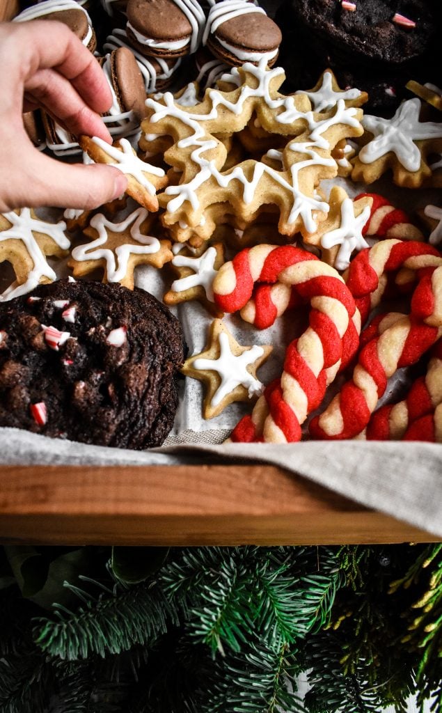 a hand reaching into a Christmas treat box to pick up a snowflake cookie