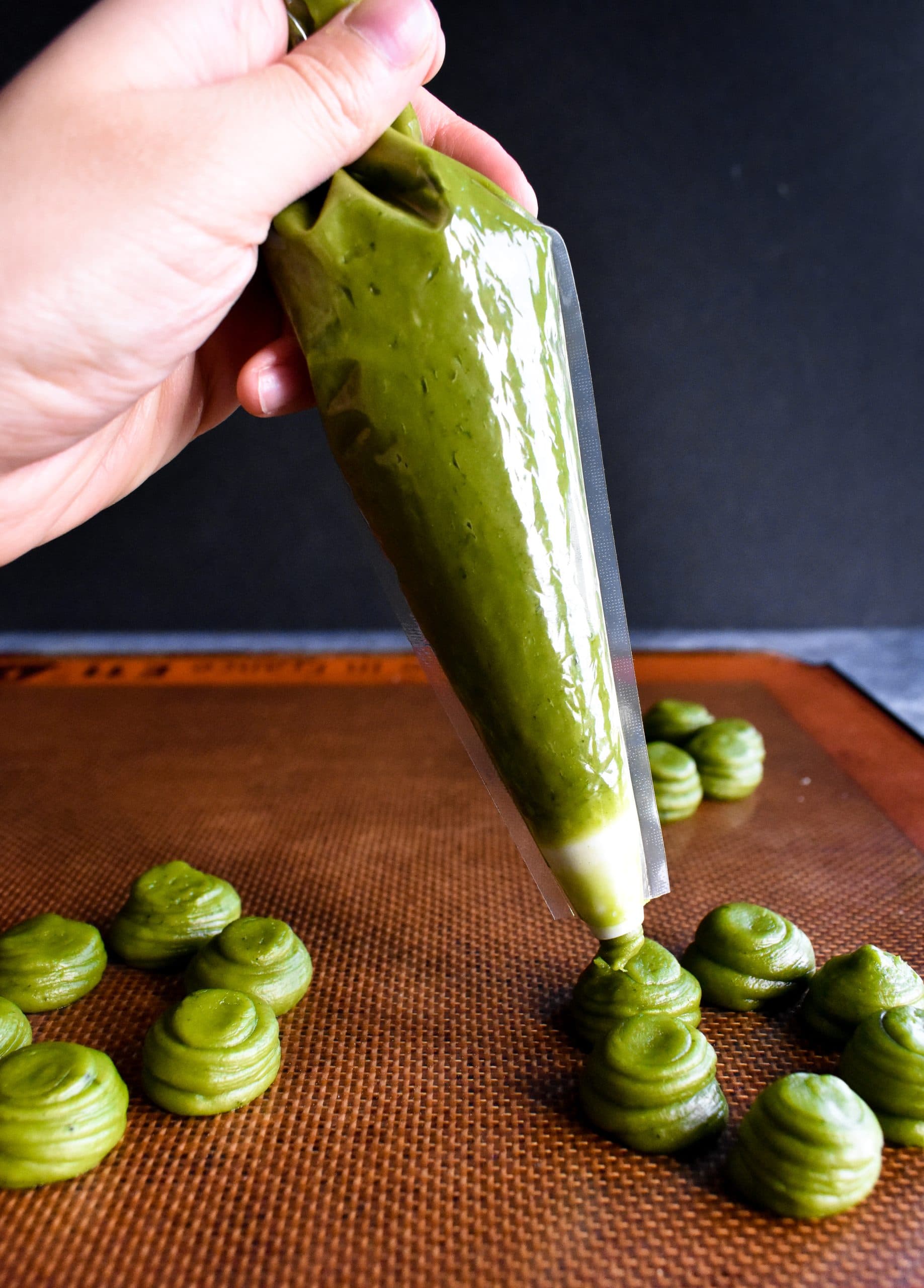Green mochi donut batter being piped onto a baking sheet with a clear piping bag.