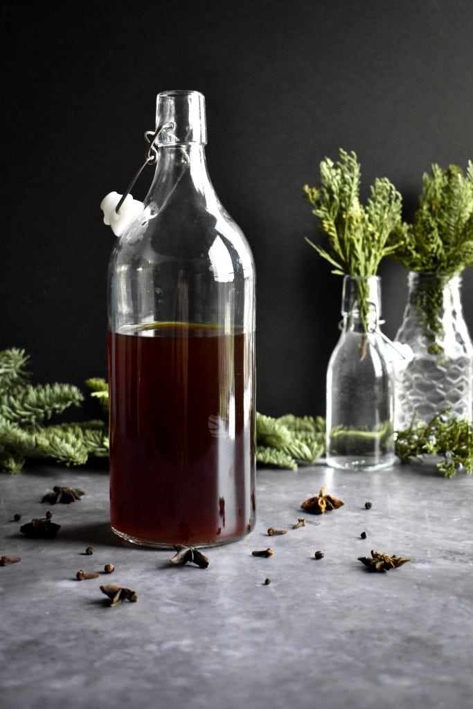 A large glass bottle filled with a dark liquid on a table next to evergreen sprigs. 