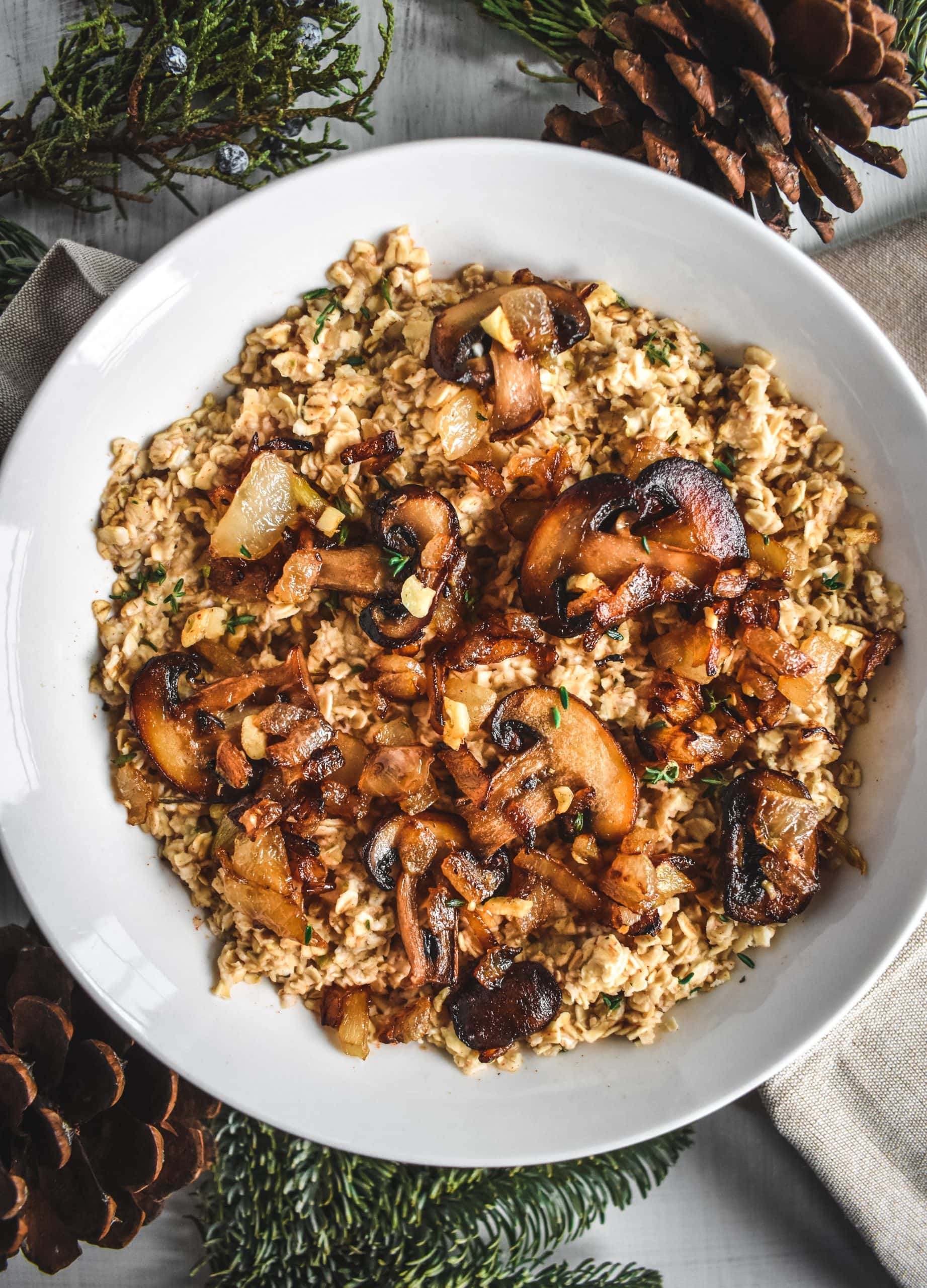 A white bowl full of creamy oatmeal with caramelized onions & mushrooms next to pinecones and evergreen sprigs.