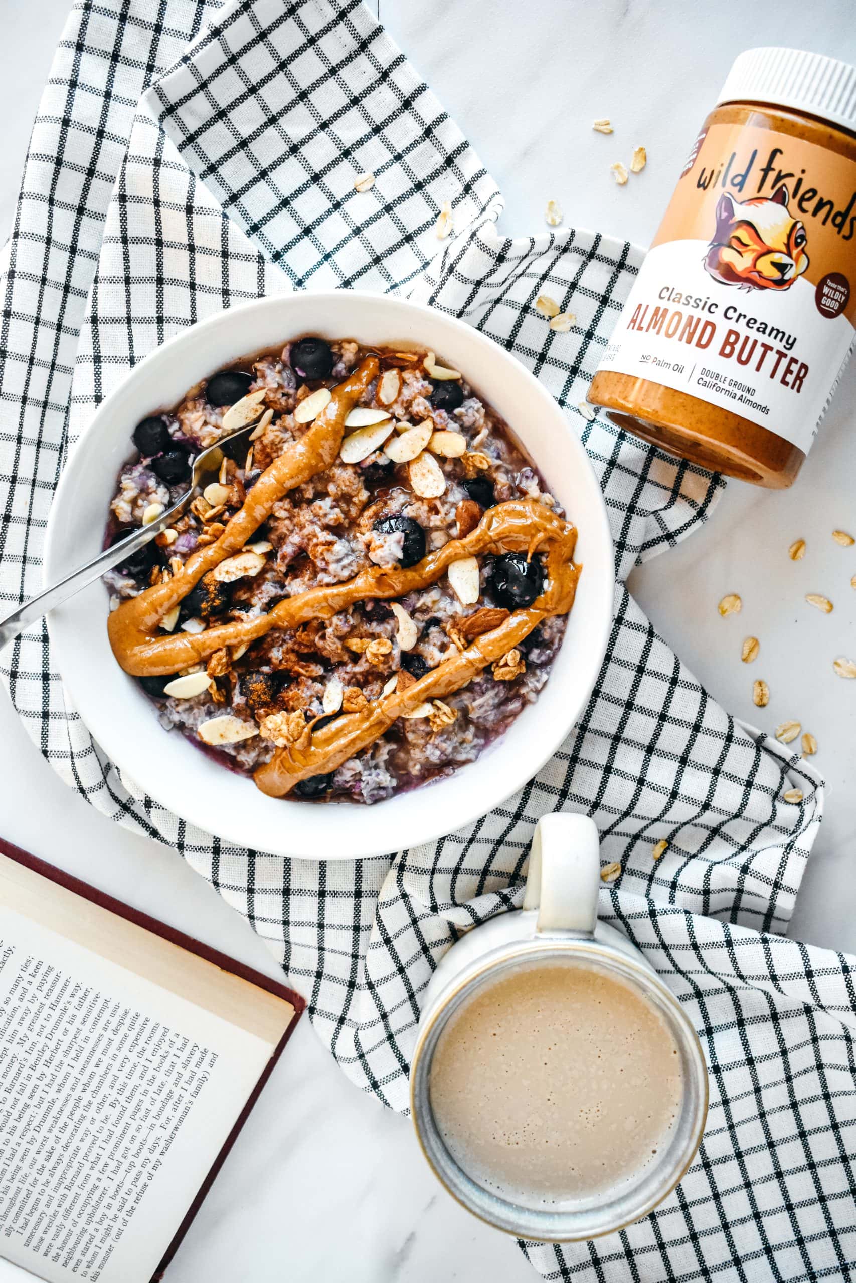 A jar of nut butter next to a bowl of blueberry oatmeal and a mug of coffee.