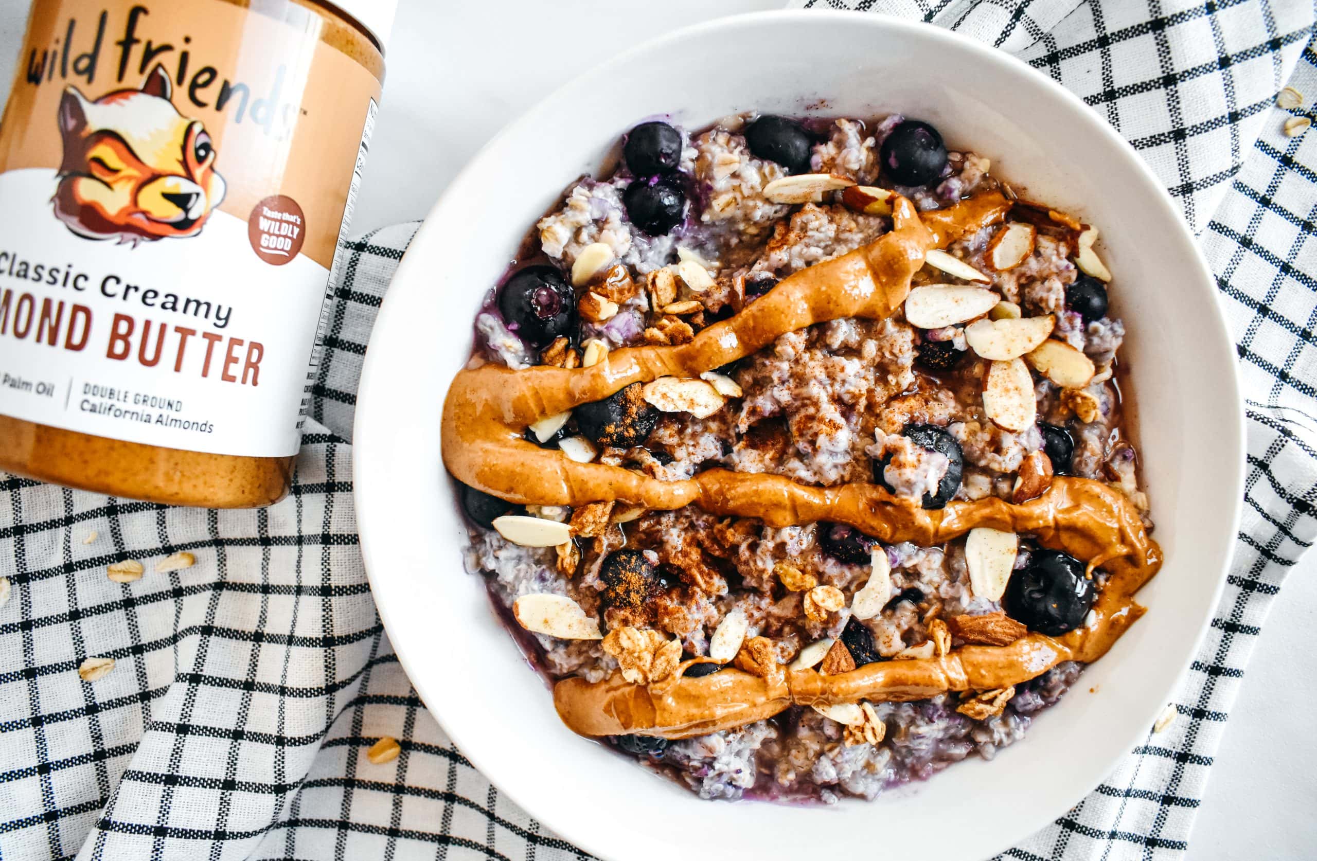 Almond butter drizzled onto a white bowl filled with oatmeal, sliced almonds, and blueberries.
