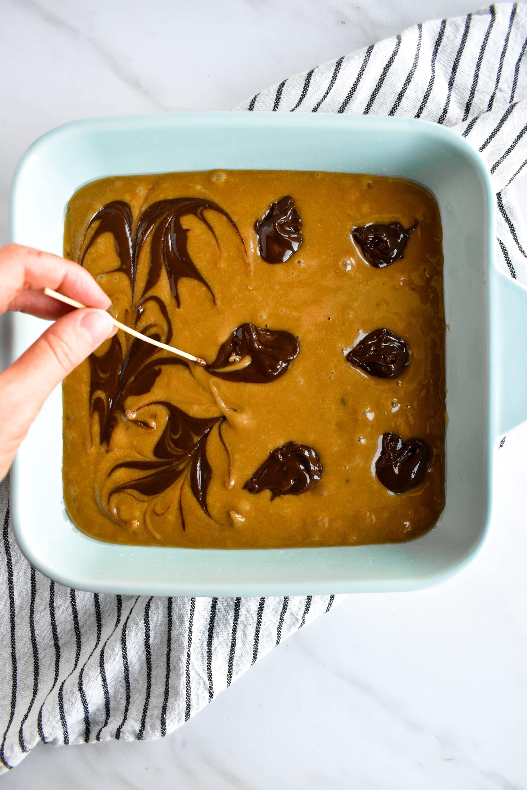 A hand swirling chocolate hazelnut spread into blondie batter in a square baking pan.