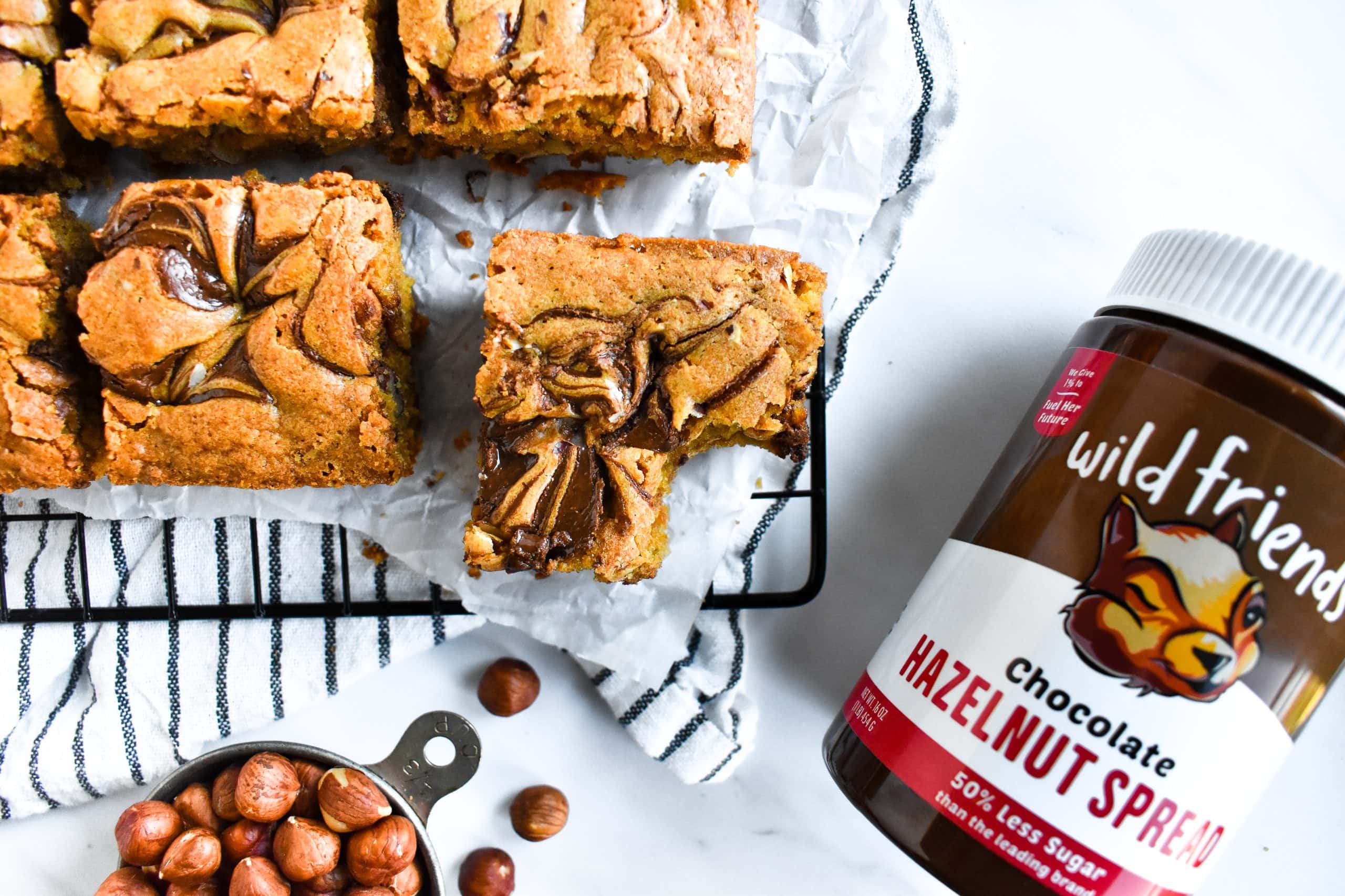 A half-eaten blondie bar sitting on a cooling rack next to a jar of chocolate hazelnut spread.