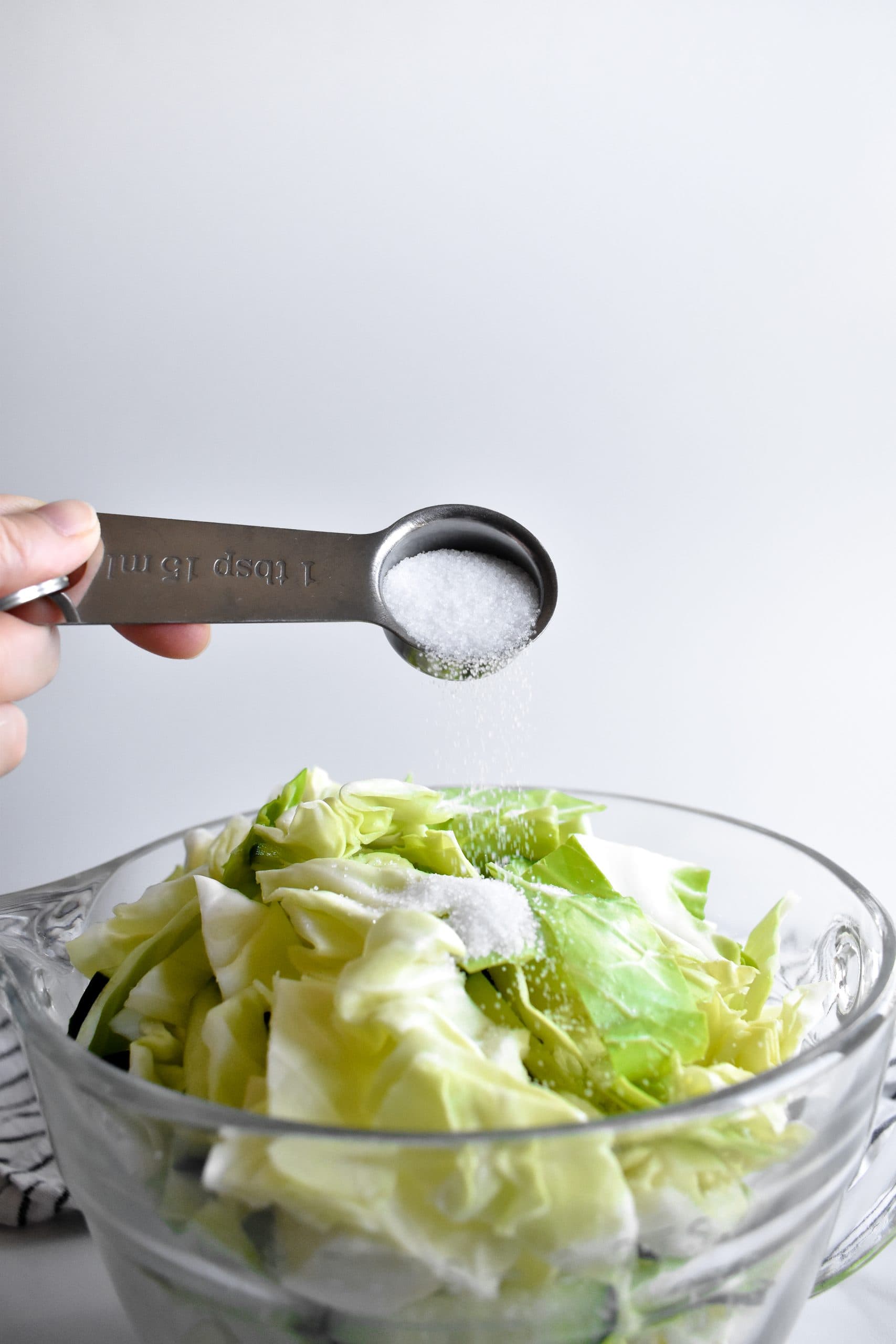 A hand pouring salt from a tablespoon onto a bowl of chopped cabbage and cucumbers.