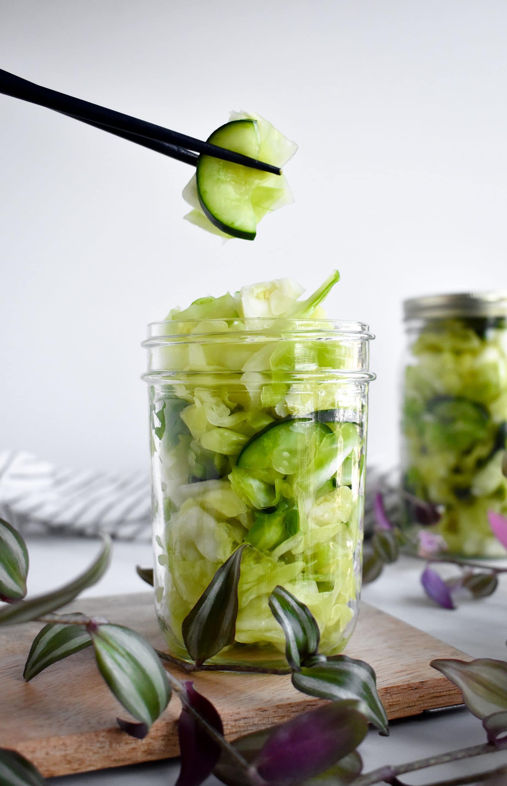 A pair of chopsticks holding fermented cabbage and cucumber over a glass jar. 