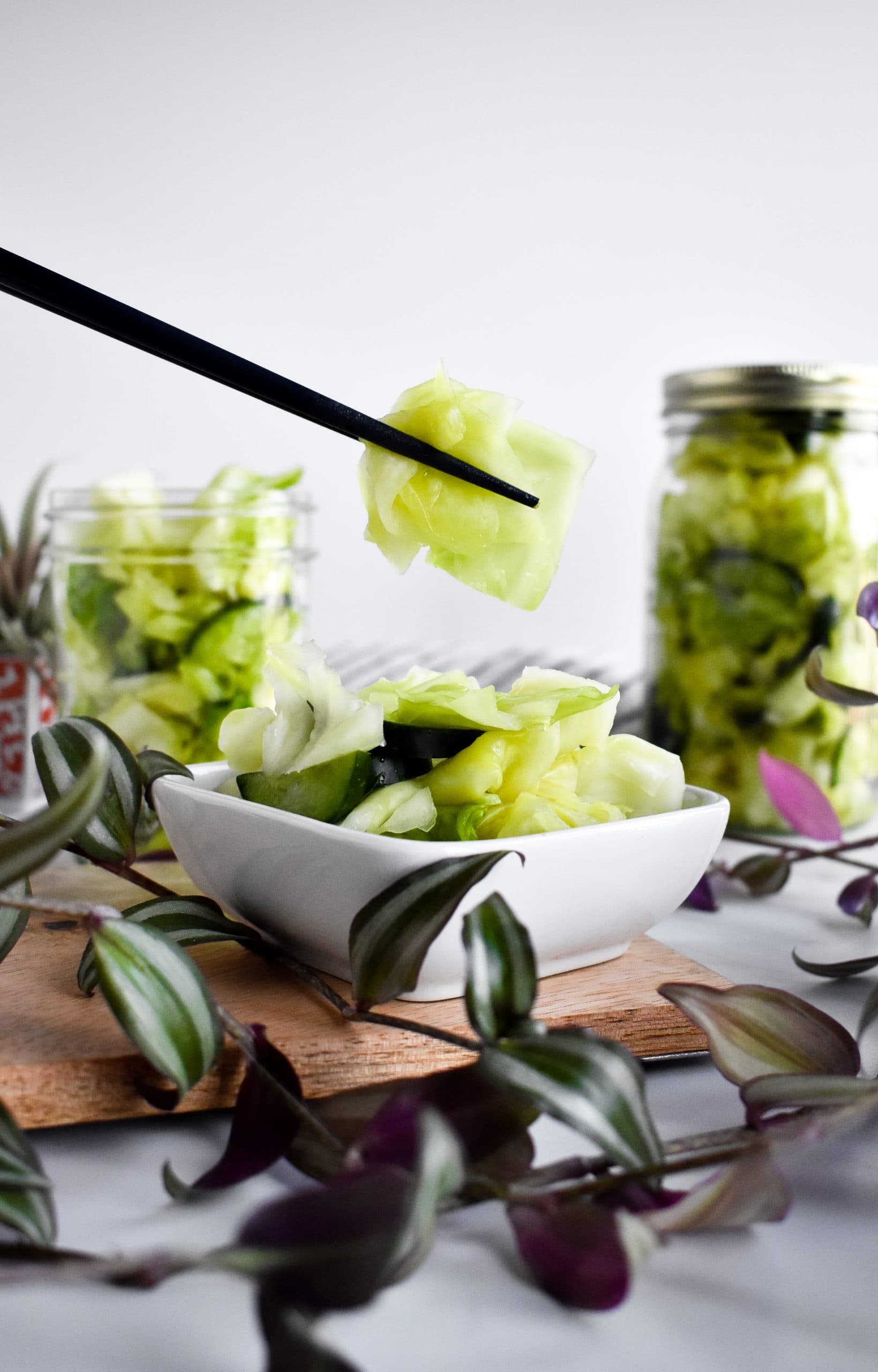 A pair of black chopsticks holding a bite of green mild kimchi next to two other glass jars filled with kimchi.