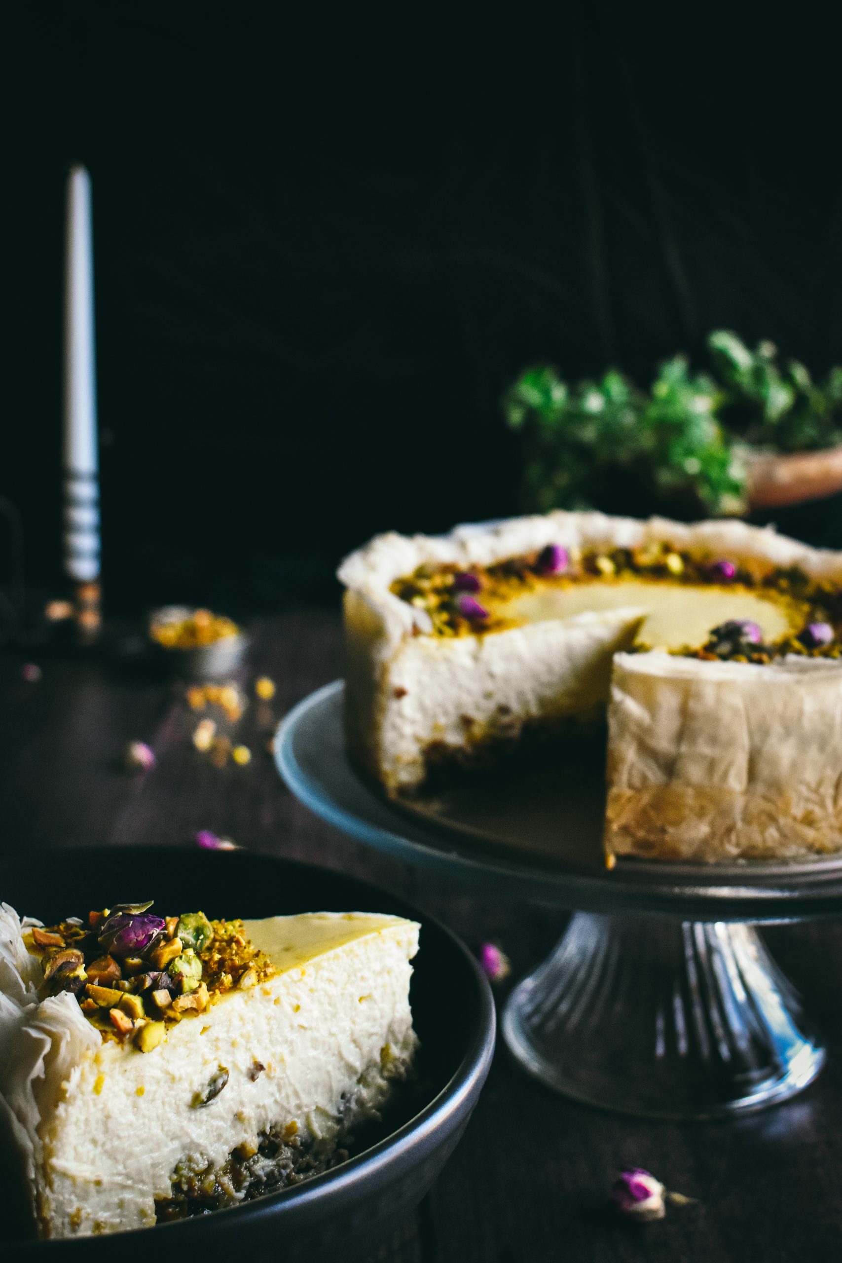 Side view of a slice of cheesecake on a black plate next to the rest of the cheesecake on a cake stand.
