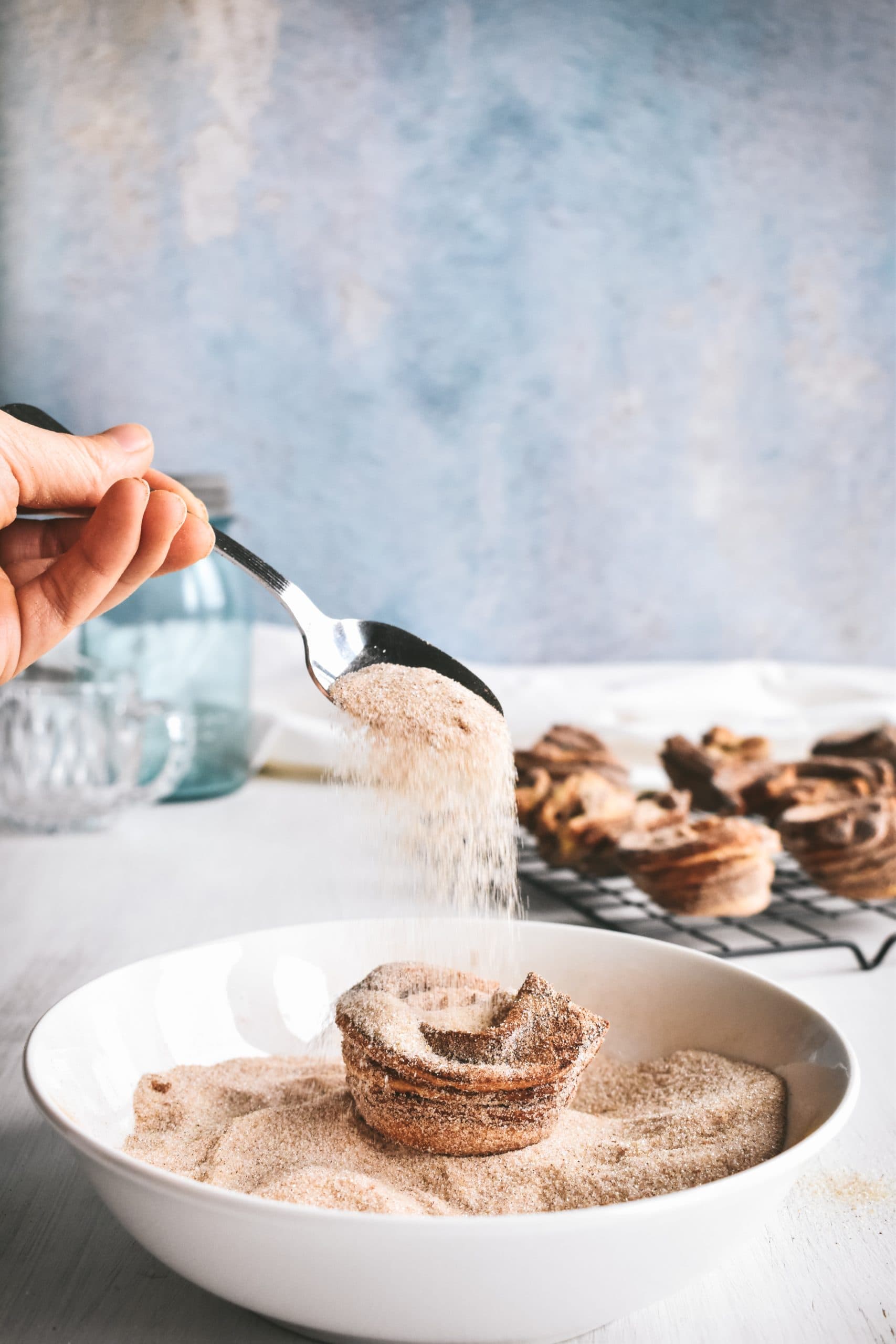 Cinnamon sugar falling off a spoon and onto a baked cruffin sitting in a shallow white bowl.