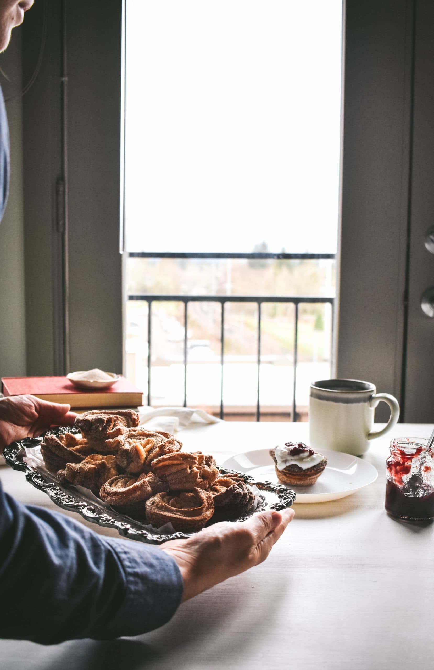 A girl setting down a silver tray of sweet breakfast baked treats on a white table. 