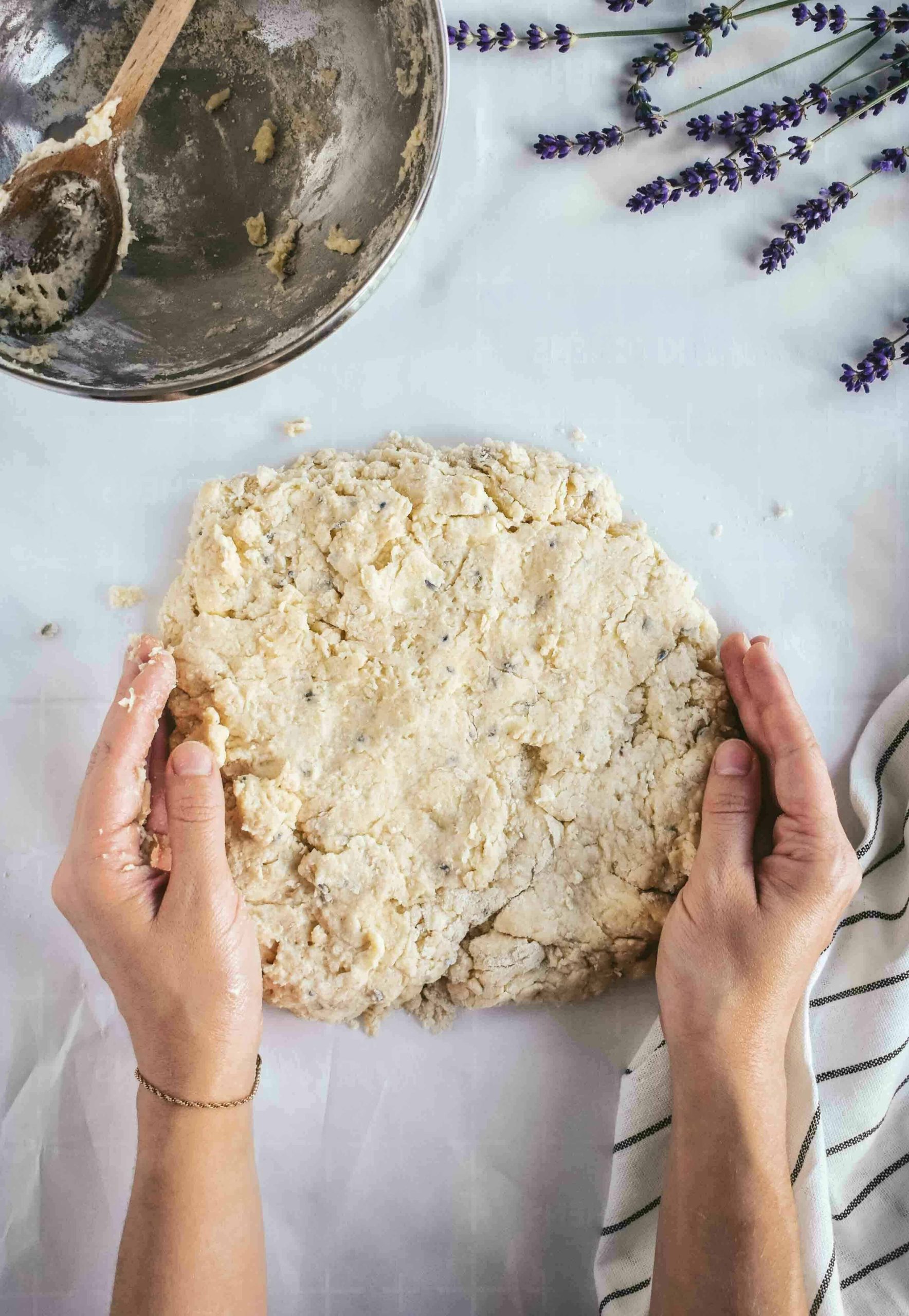 Two hands shaping scone dough into a large flat disc on a white surface.