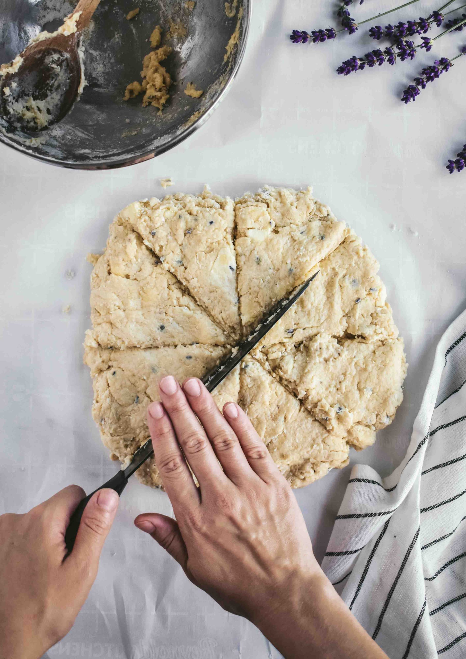 Two hands cutting a disc of scone dough into triangles with a large knife on a light surface.