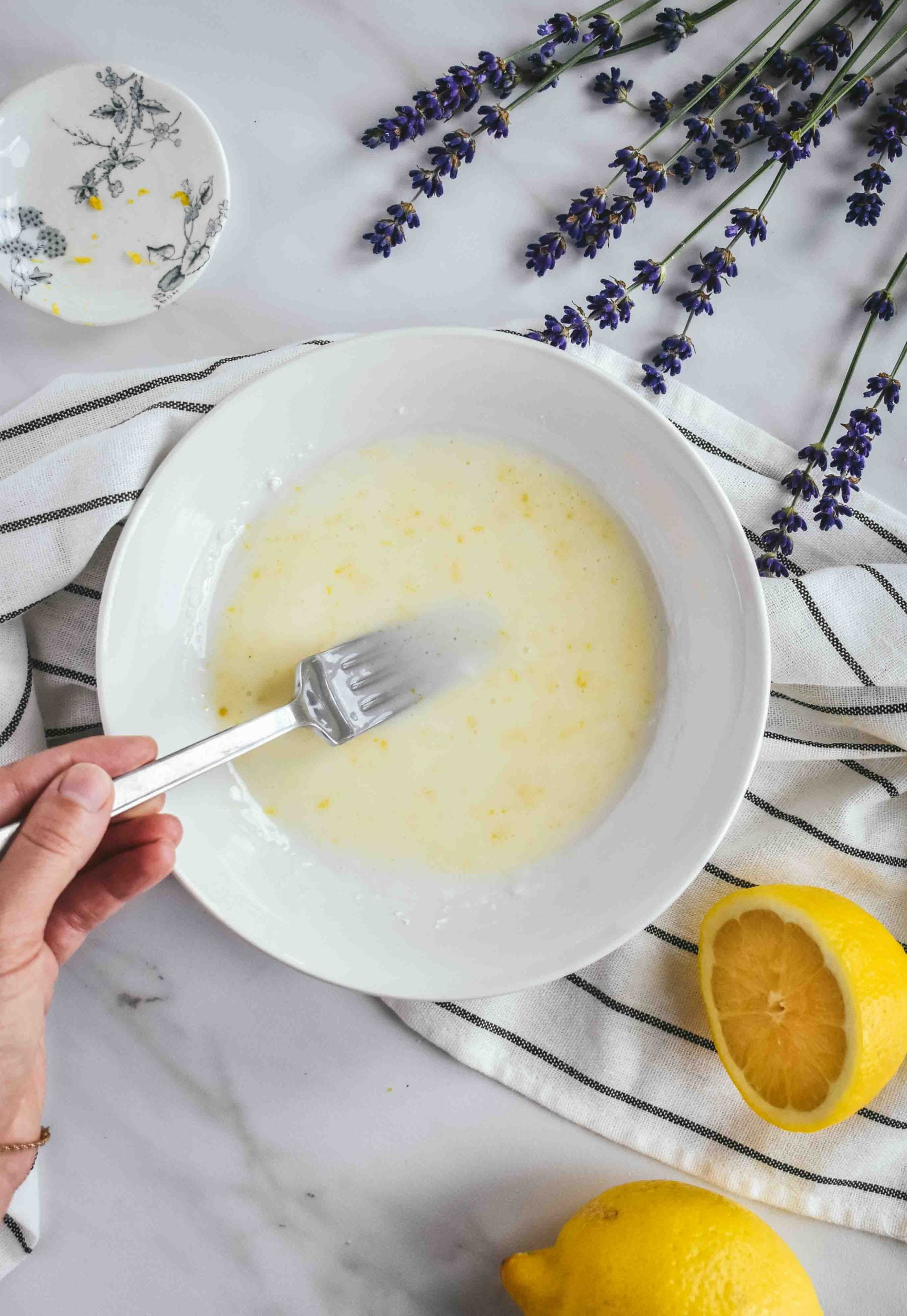 A hand mixing lemon glaze in a white bowl with a fork on a white and black linen.