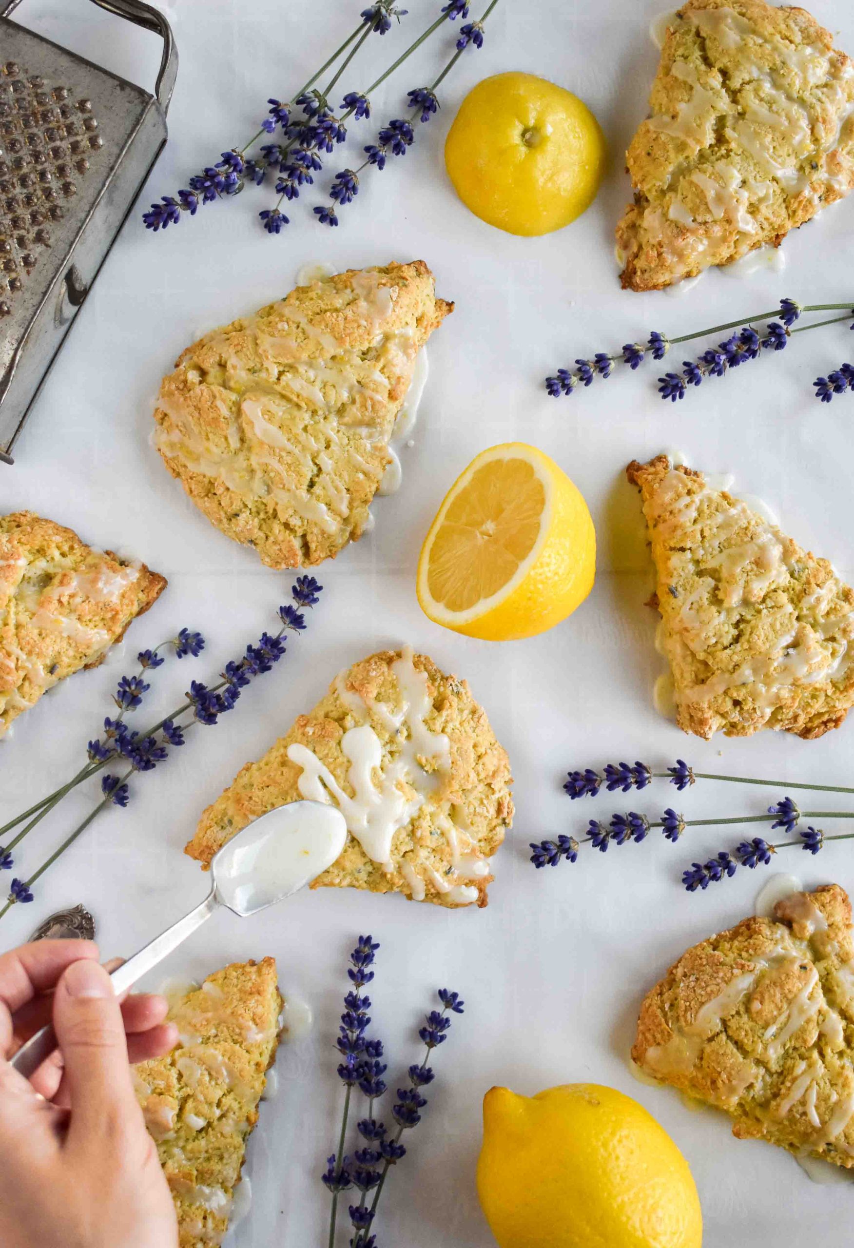 A hand drizzling white glaze onto baked lemon lavender scones next to fresh halved lemons and sprigs of lavender.
