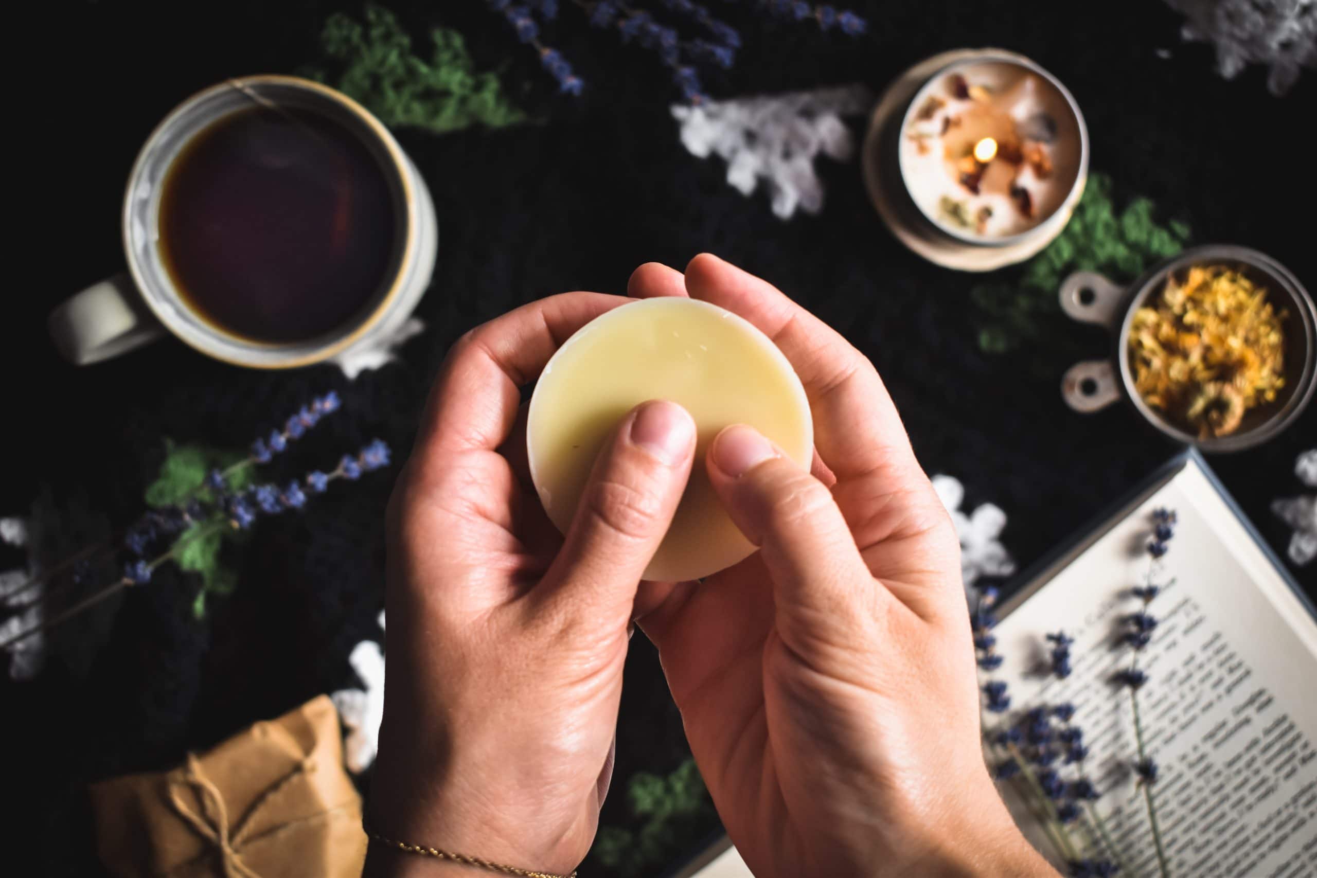 Two hands holding a Homemade Lotion Bar with a book and a candle.