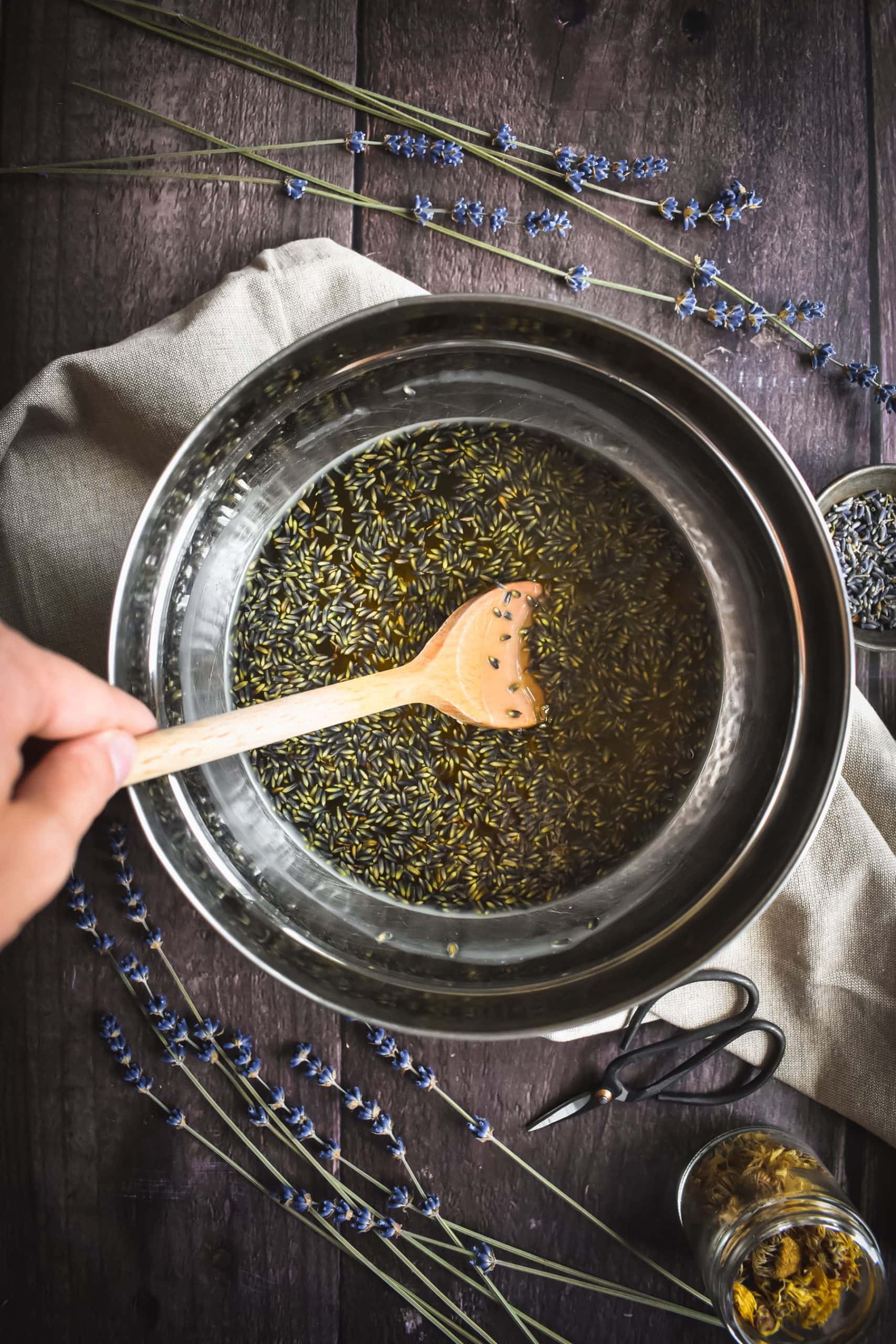 A hand stirring a wooden spoon in a metal bowl of lavender flowers.