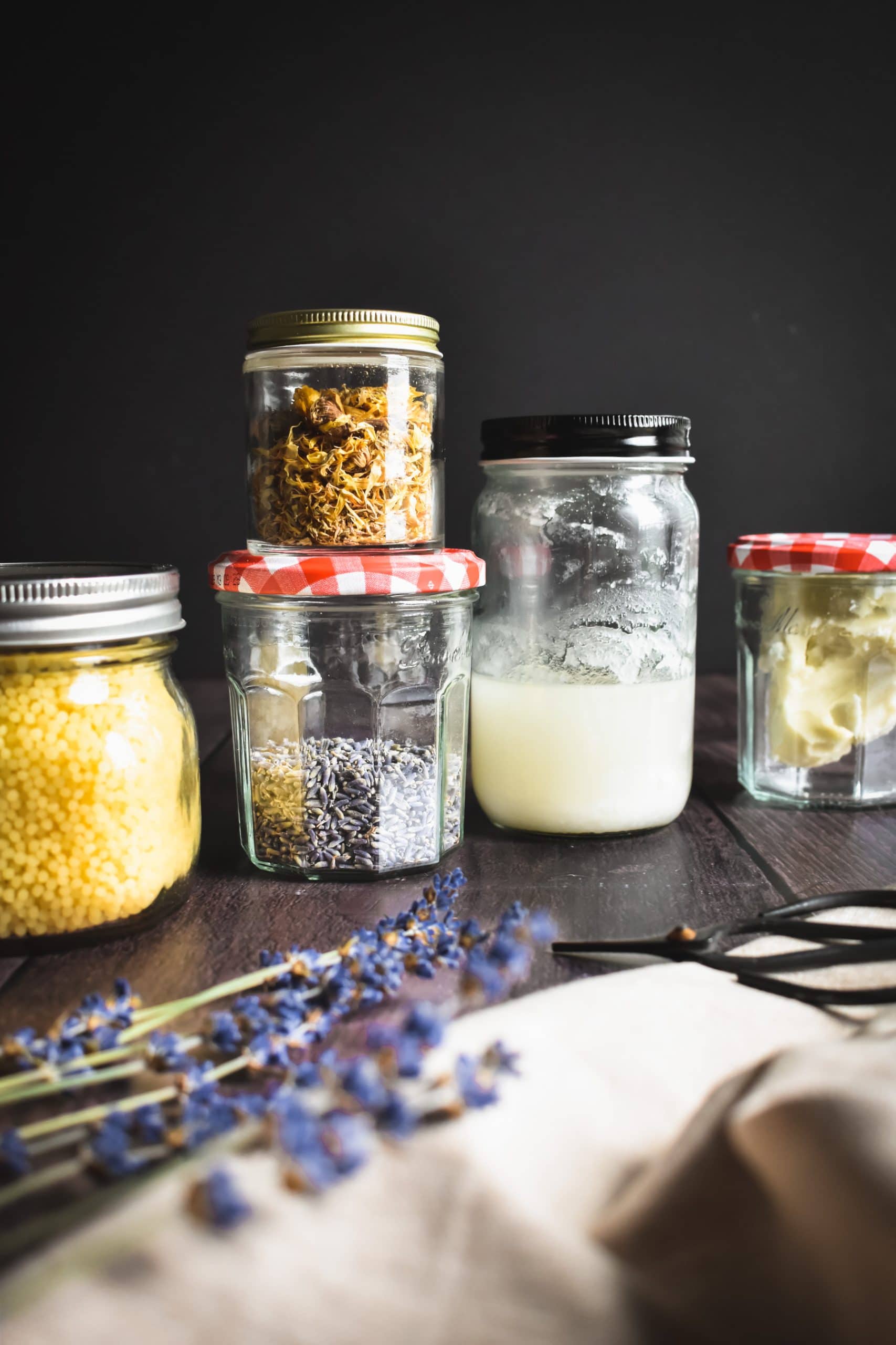 Glass jars of cocoa butter, lavender sprigs, beeswax, and coconut oil on a table.