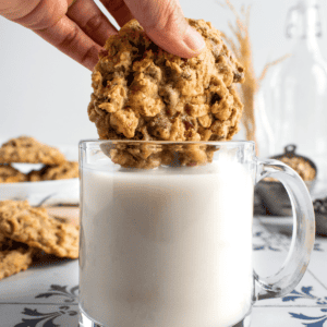 A hand dunking a chunky oatmeal cookie into a glass mug of milk.