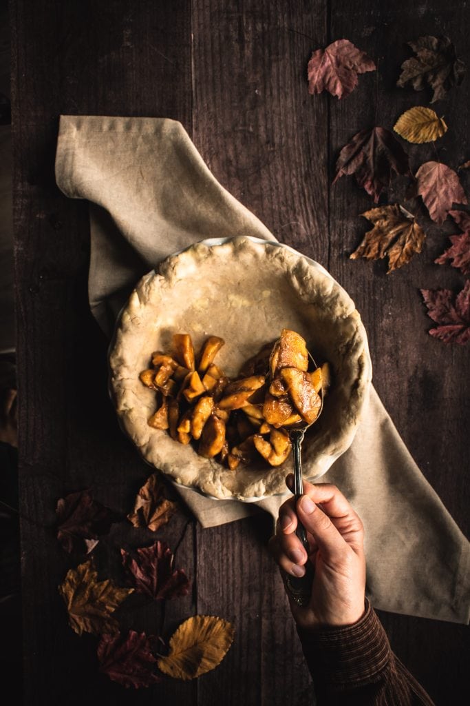 A hand placing a spoonful of apple pie filling into a pie crust.