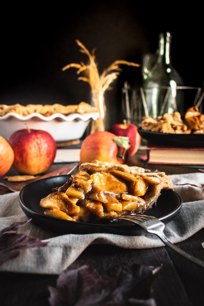Close up of a slice of German apple pie on a black plate next to whole apples.