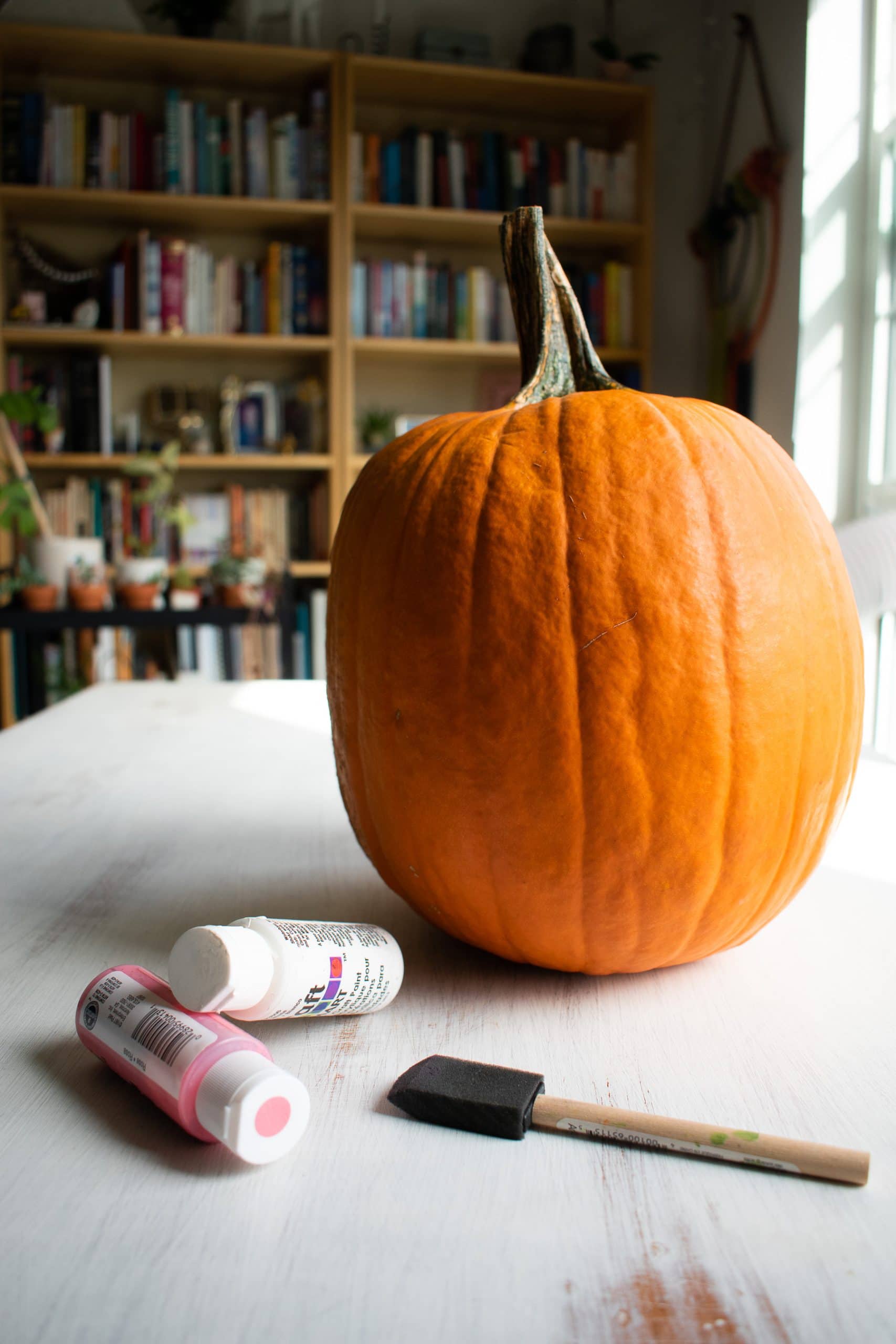 A big orange pumpkin next to bottles of white and pink paint.