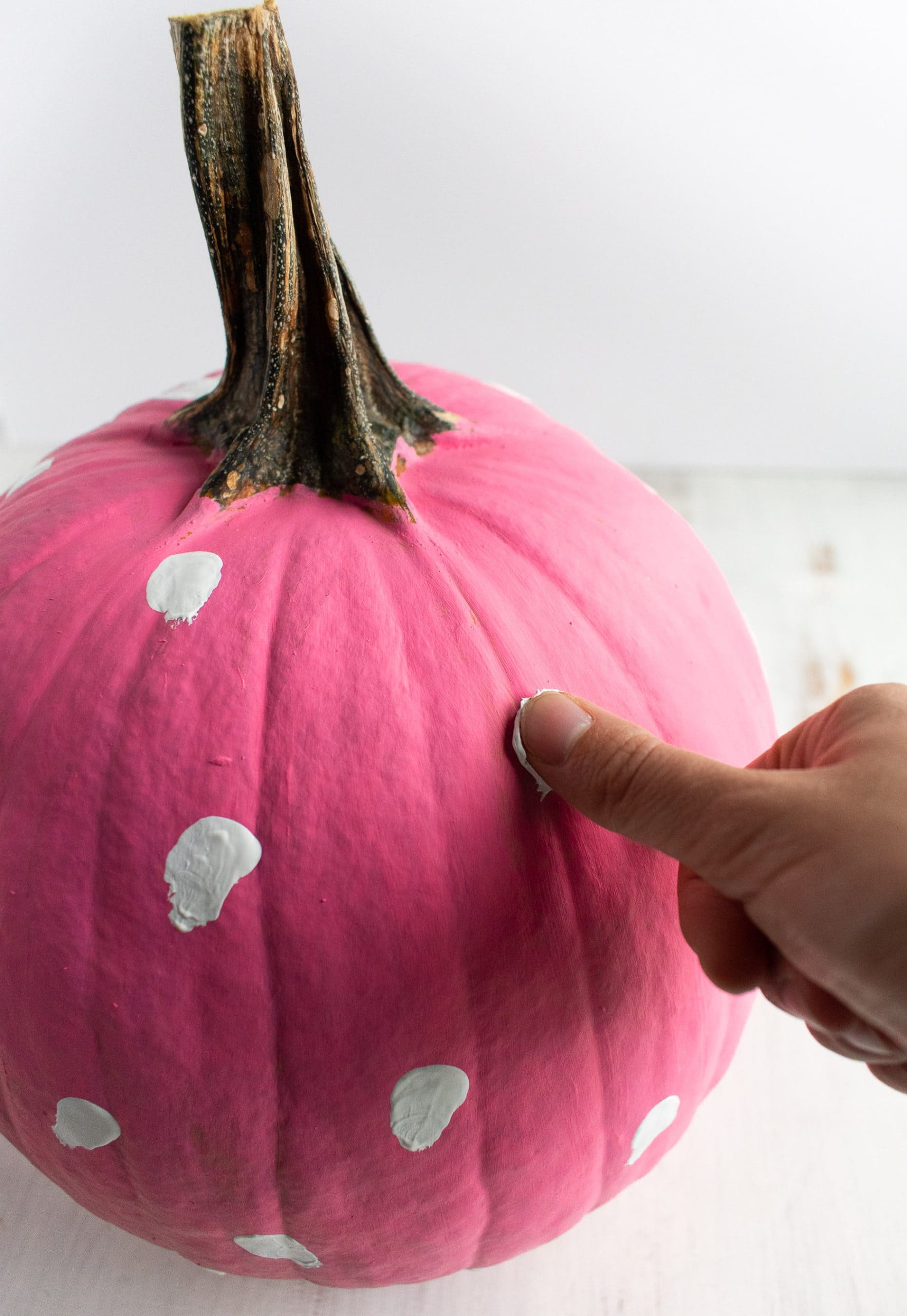 A girl's thumb pressing white paint onto a pink pumpkin.