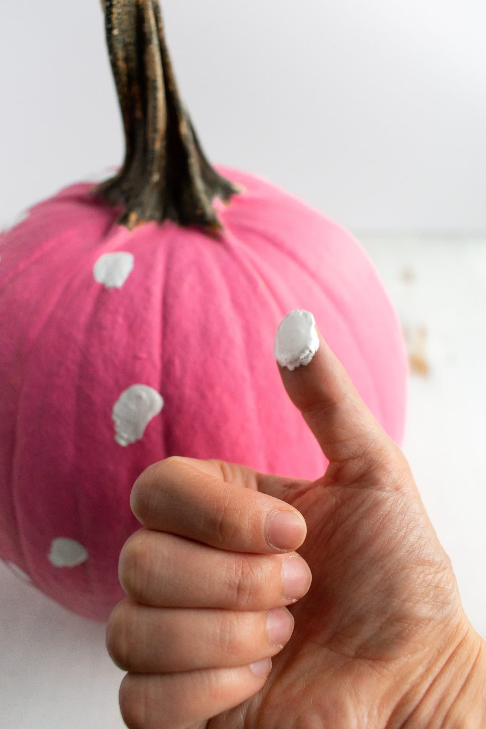 A girl's thumb covered with white paint to press onto a pink painted pumpkin.
