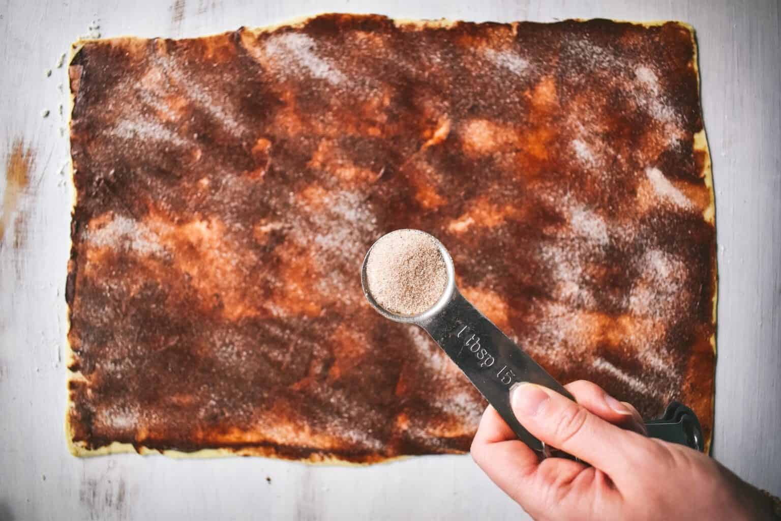 A hand sprinkling a spoonful of cinnamon sugar onto a sheet of puff pastry dough.