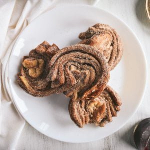 Four cruffins stacked on a white plate on a white table, coated in cinnamon sugar.