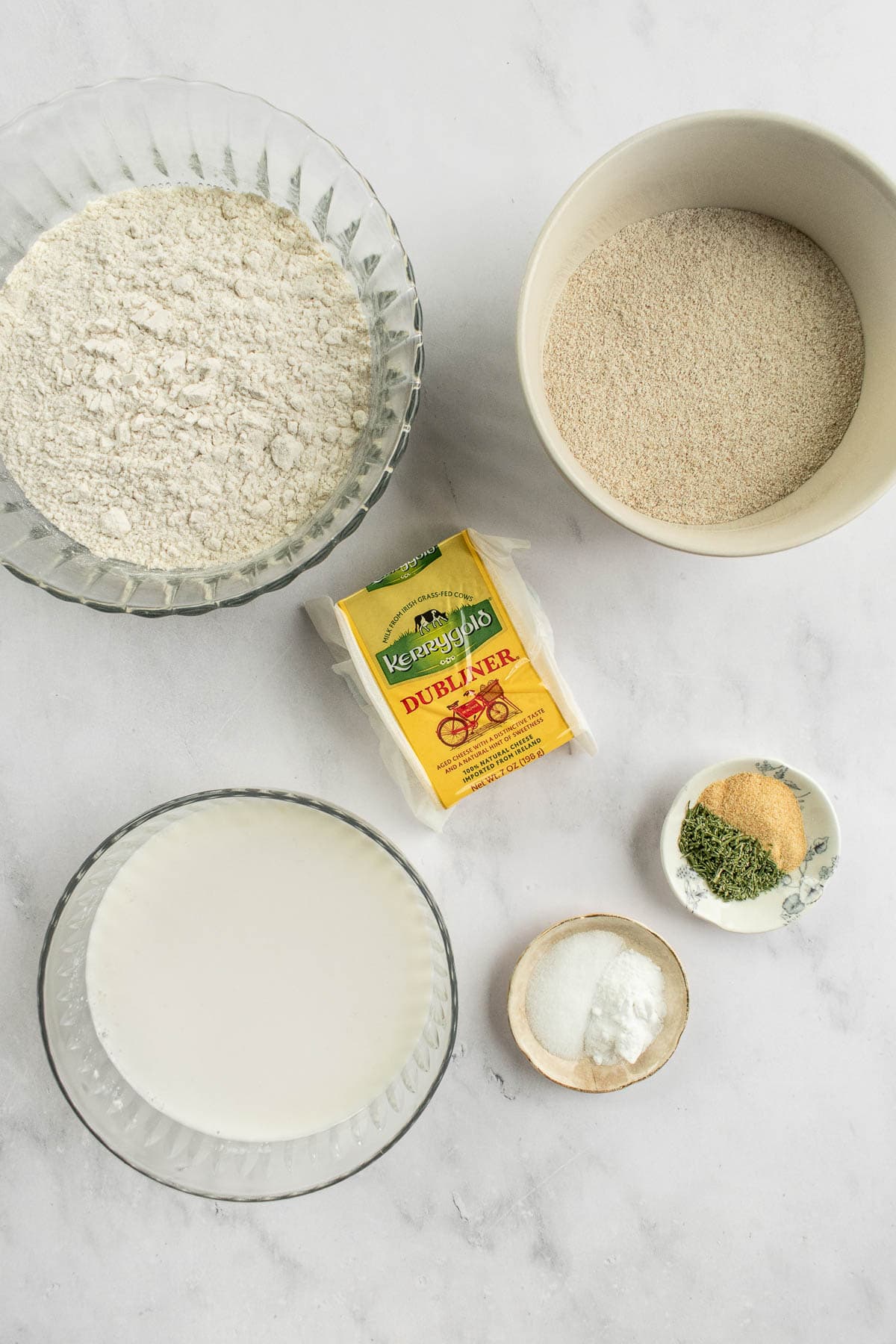 Bowls of white flour, rye flour, salt, dried herbs and sides, and buttermilk on a table next to a package of Irish cheese.