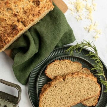 Two slices of hearty soda bread on a metal plate next to the rest of the loaf on a green linen.