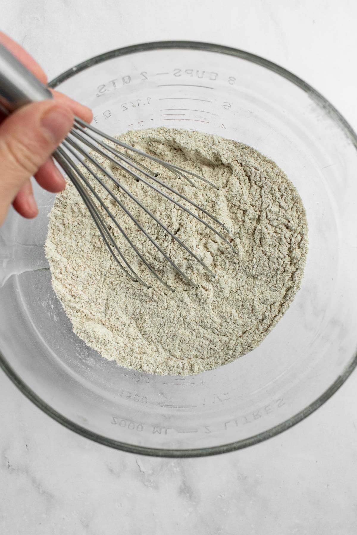 A hand whisking rye flour, white flour, spices, dried herbs, salt, and yeast in a glass bowl.