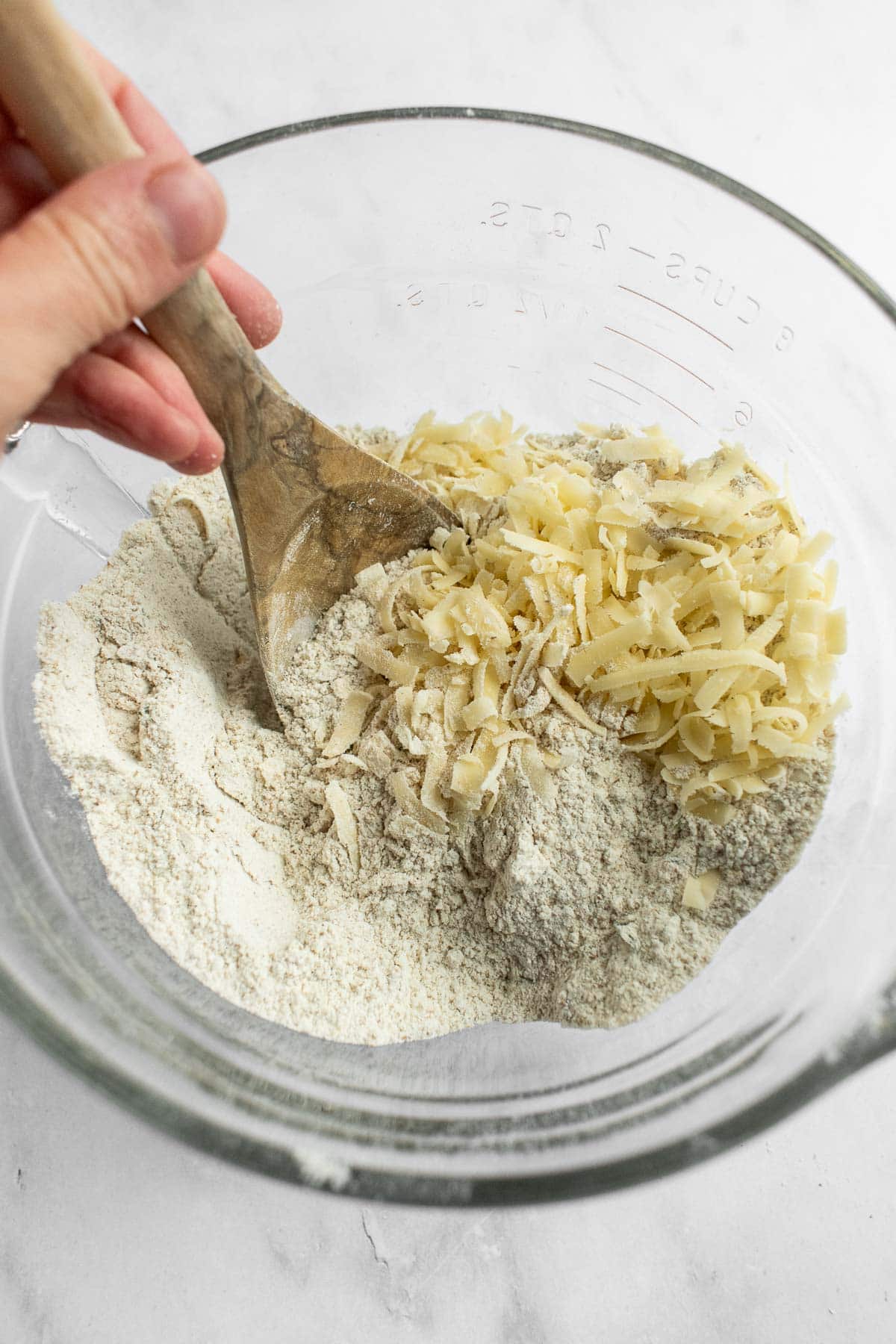 A hand stirring shredded cheese into a bowl of flour with a wooden spoon.