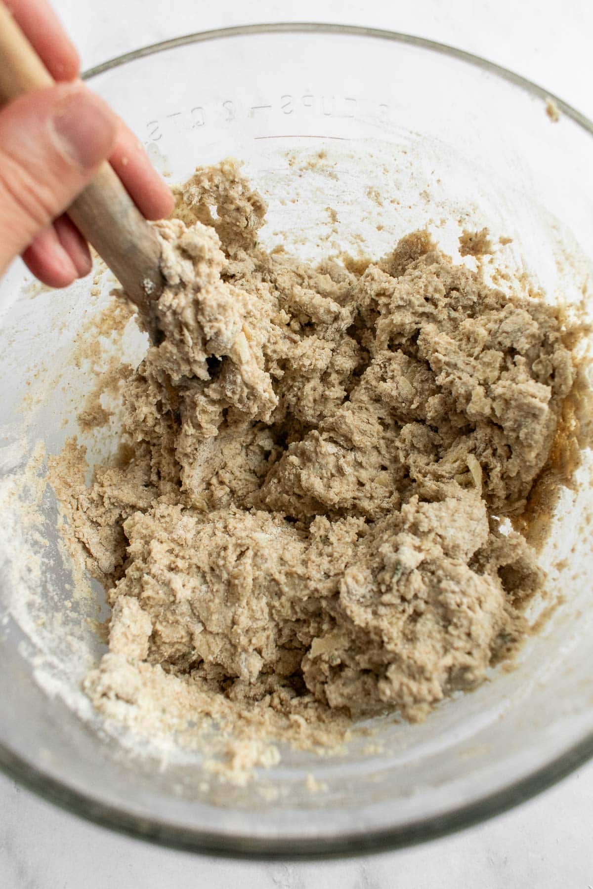 A hand mixing really thick bread dough with dried herbs and shredded cheese with a wooden spoon in a glass bowl.