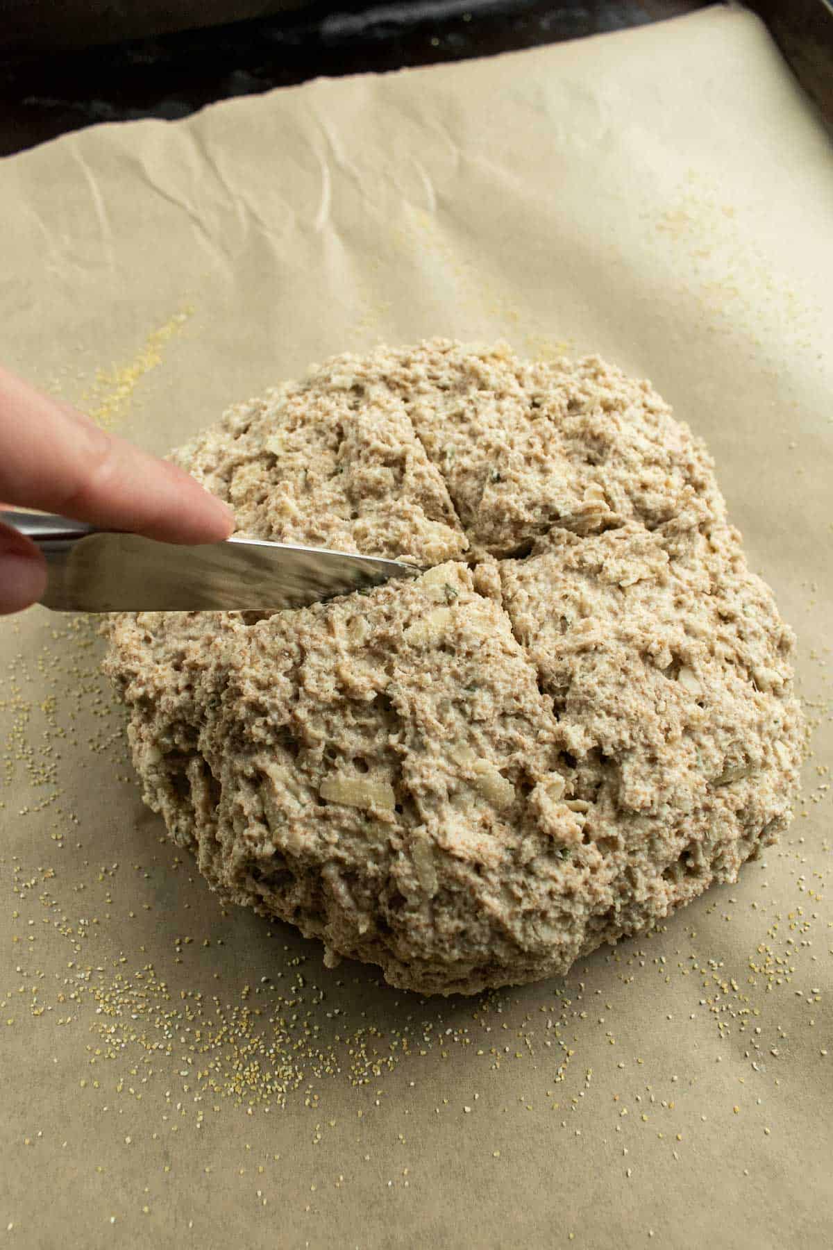 A hand scoring a loaf of soda bread dough with a knife on a baking sheet lined with parchment paper.