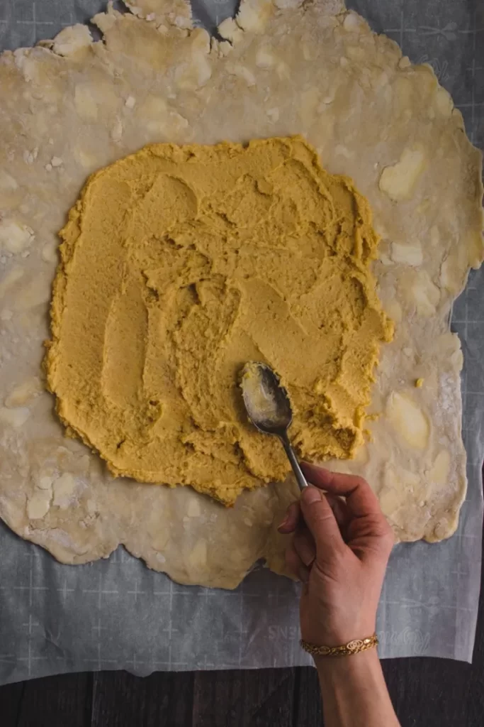 Hummus being spread on the pie dough with a spoon.