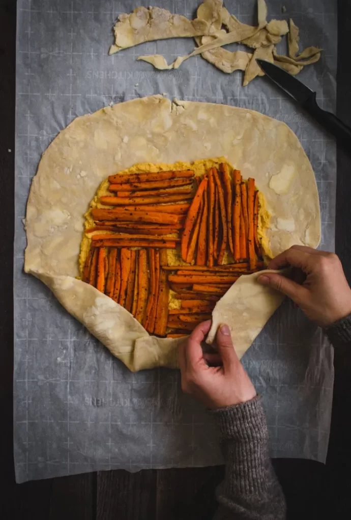 Two hands folding pie crust over roasted carrots to form a galette.