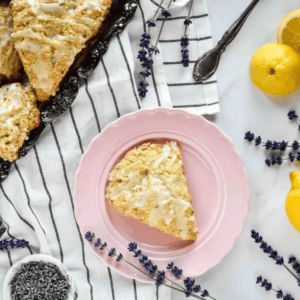 A lavender scone topped with lemon glaze on a pink plate next to whole lemons and lavender sprigs.