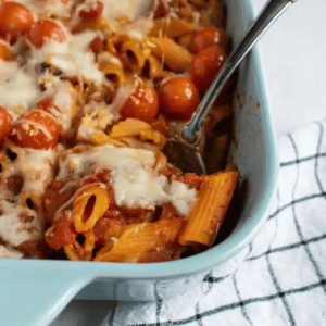 A baking dish filled with cooked penne pasta in red sauce with cherry tomatoes and melted cheese.