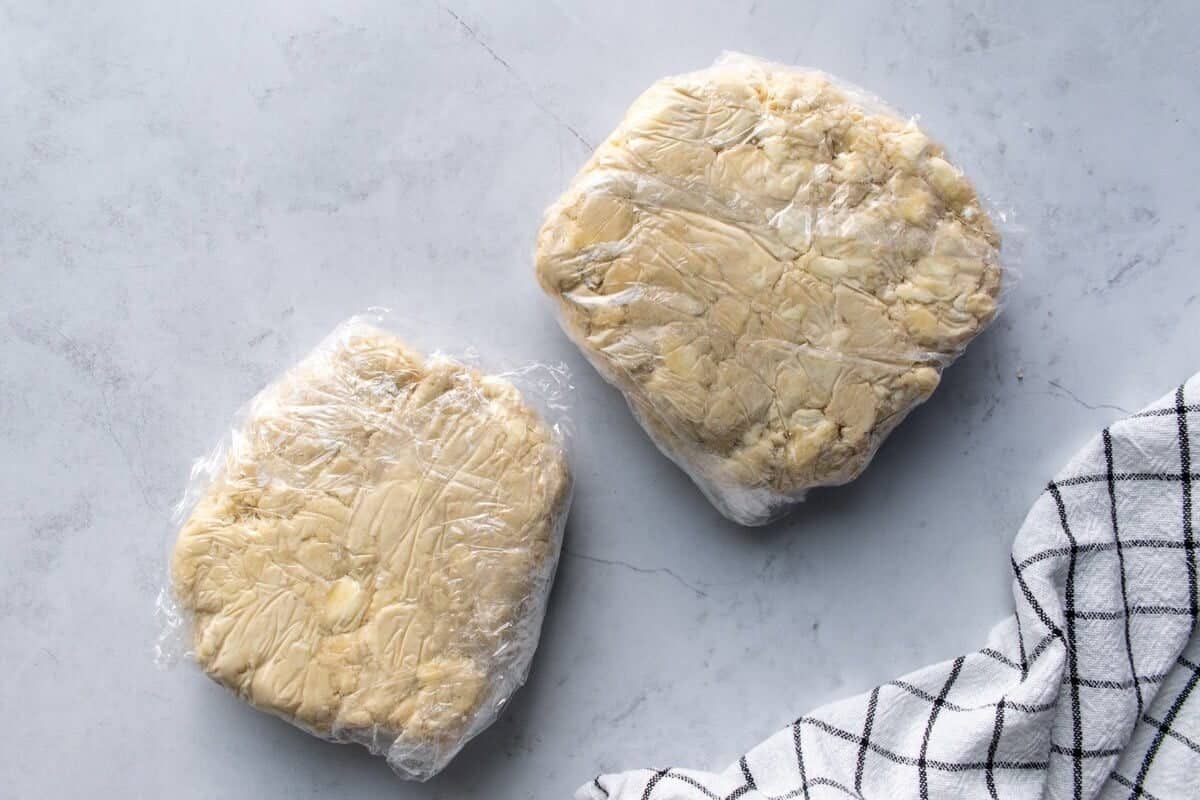 Two discs of pie dough wrapped in plastic wrap on a light surface.