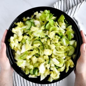 Hands holding a bowl of chopped cabbage and cucumber over a light surface.