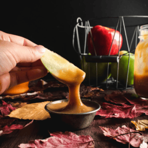 A hand holding an apple slice dripping caramel sauce into a metal dish.