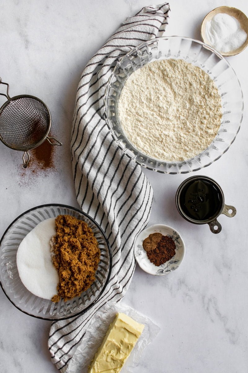 Bowls of flour, sugar, molasses, spices, and salt next to a stick of butter on a light surface.