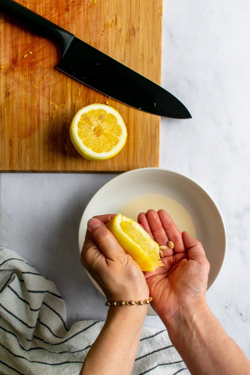 Two hands squeezing half a lemon into a shallow white bowl.